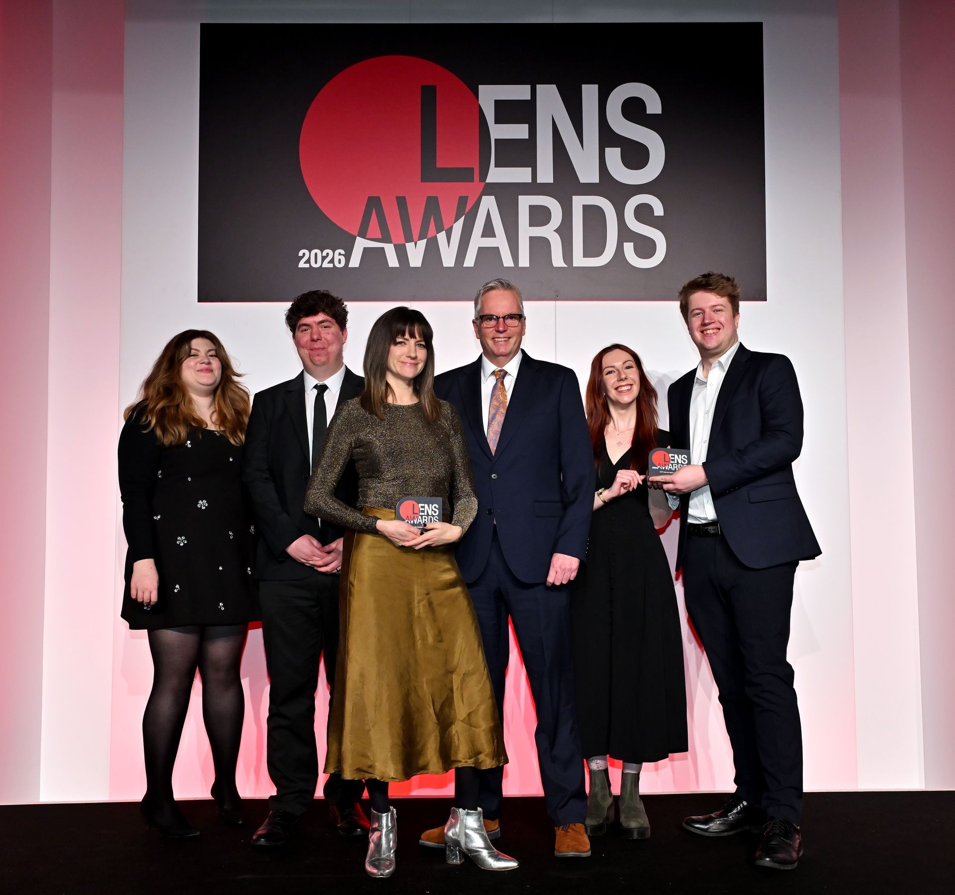 Six people pose onstage at the 2026 Lens Awards in front of a large event banner.