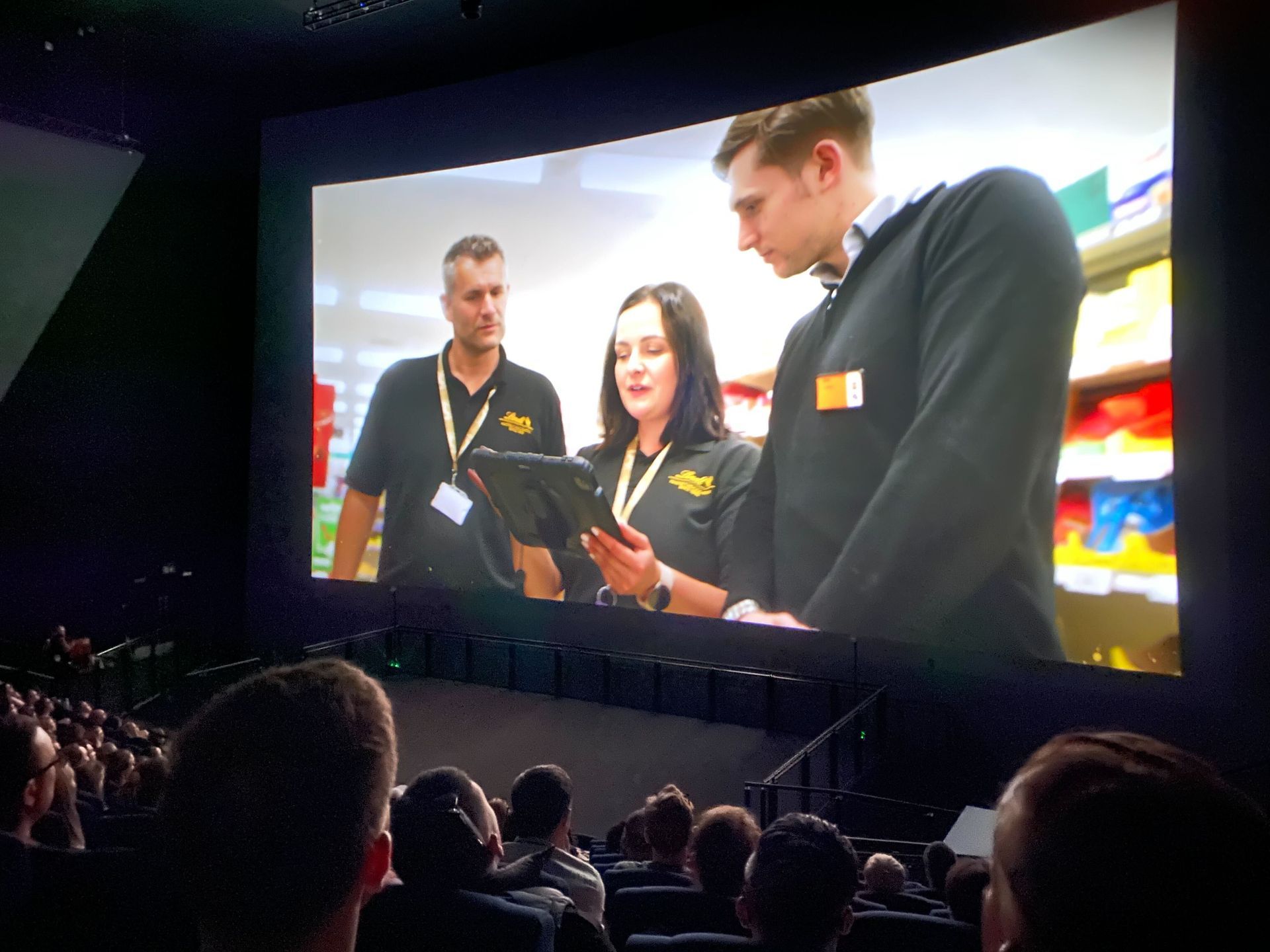 Audience watching a presentation on a large cinema screen showing three people in black shirts reviewing a tablet.