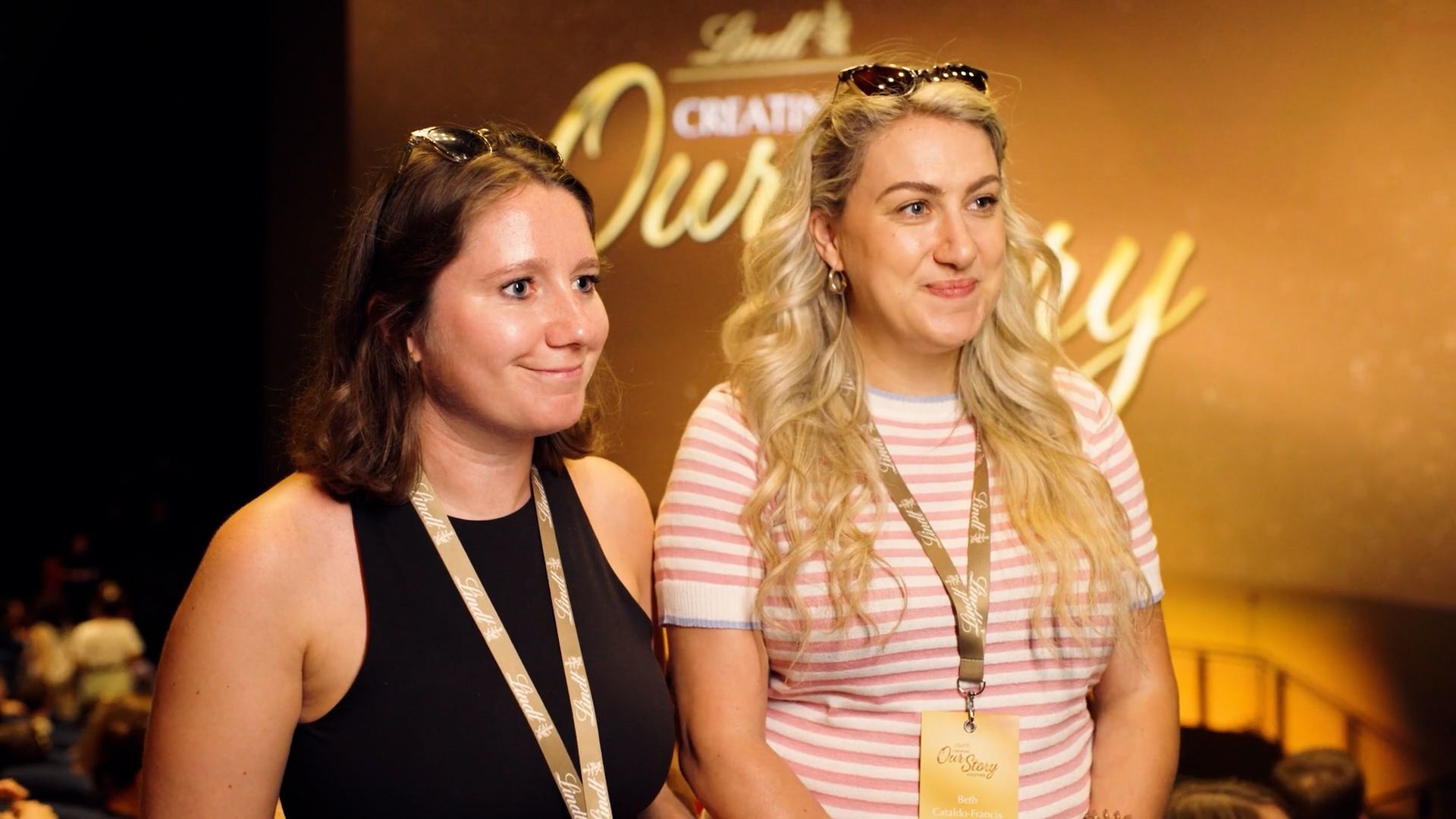 Two women standing at an indoor event, wearing lanyards, with a gold backdrop behind them