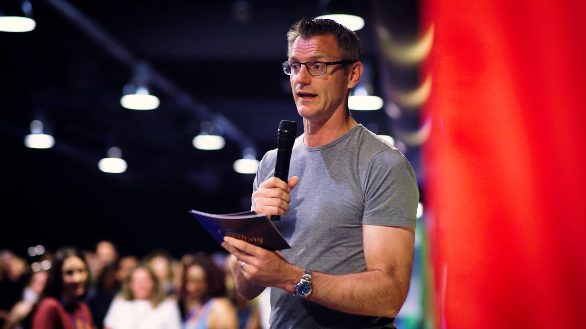 Man speaking into a microphone at a conference, with a red-lit backdrop and blurred audience behind him.