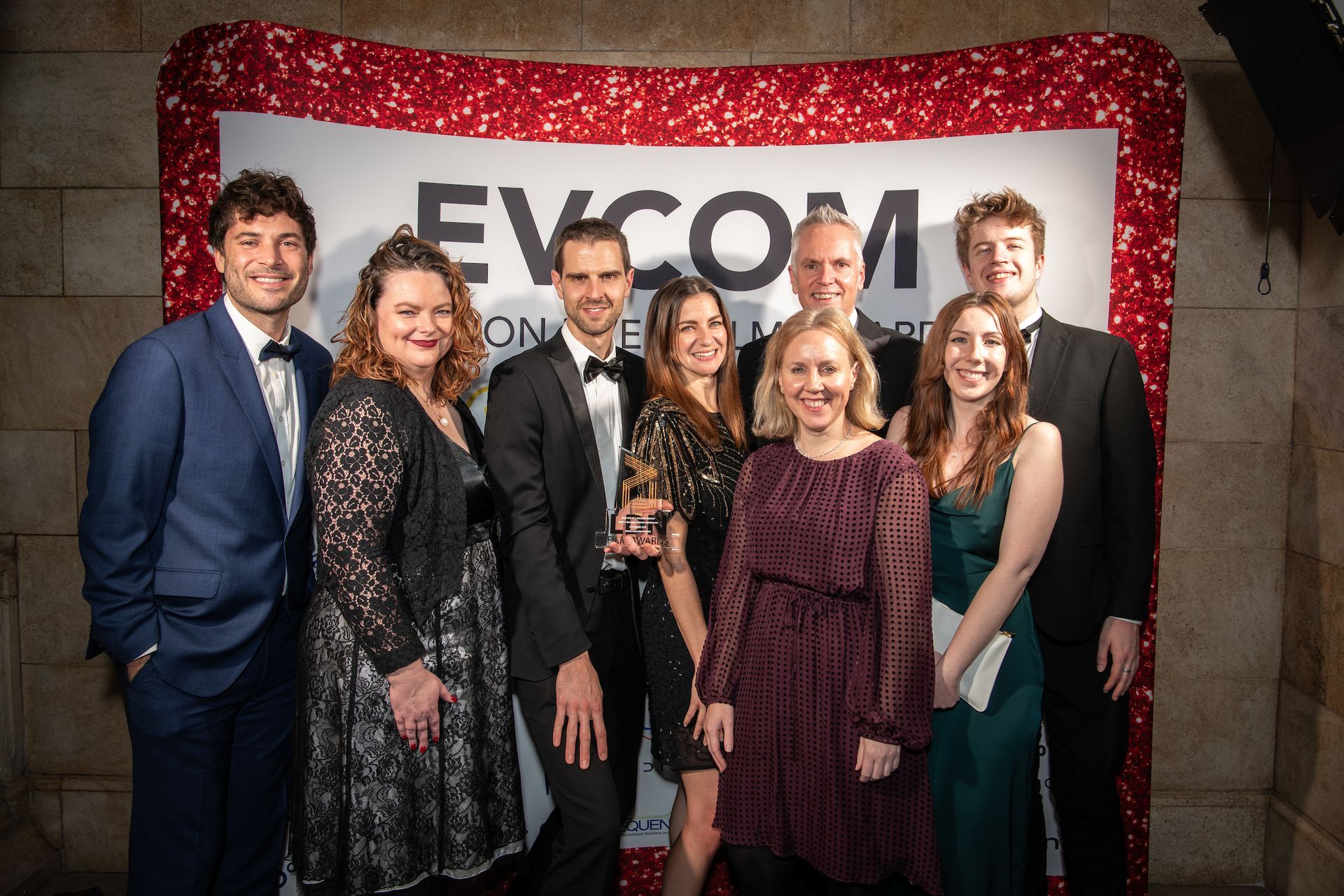 A group of people are posing for a picture in front of a sign that says evcom.
