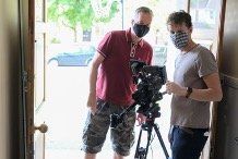 Two men wearing face masks are standing in front of a camera.