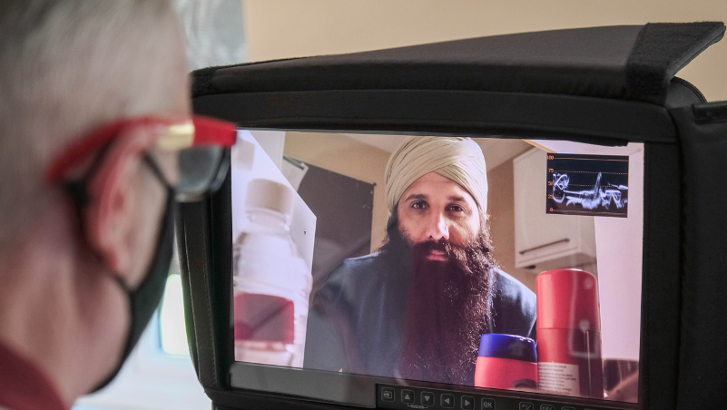 A man wearing a mask is looking at a screen with a bearded man on it.