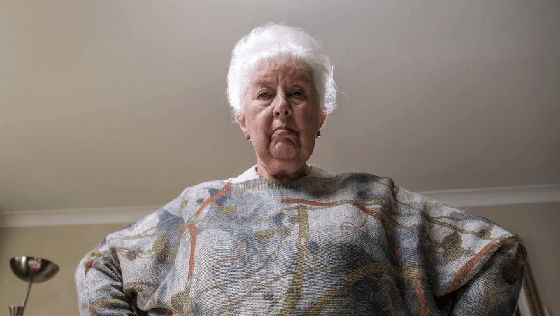 An elderly woman is standing in a living room wearing a sweater.