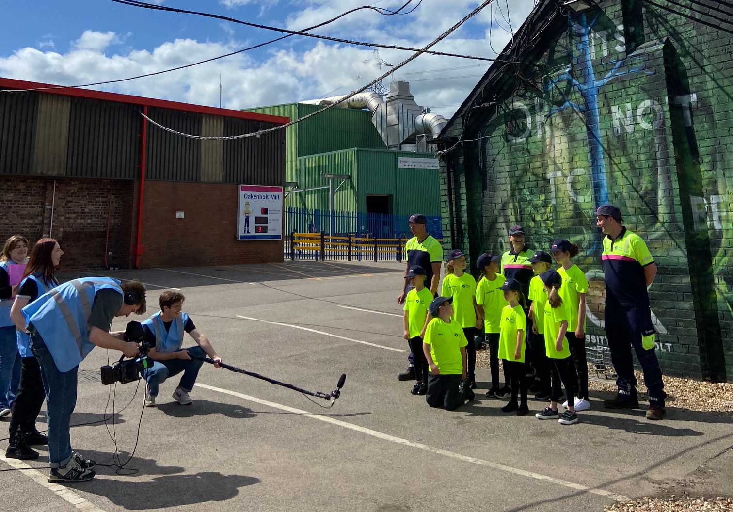 Children in neon yellow vests gathered outdoors, watching a person crouch near a long stick on the pavement.