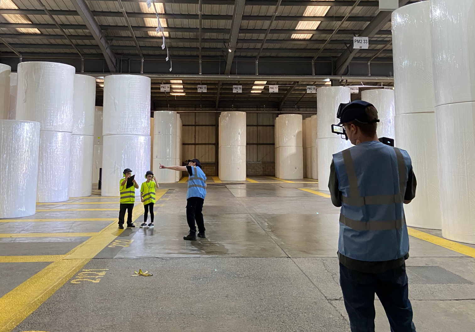 Workers in a warehouse beside large white wrapped rolls, with one person taking a photo.