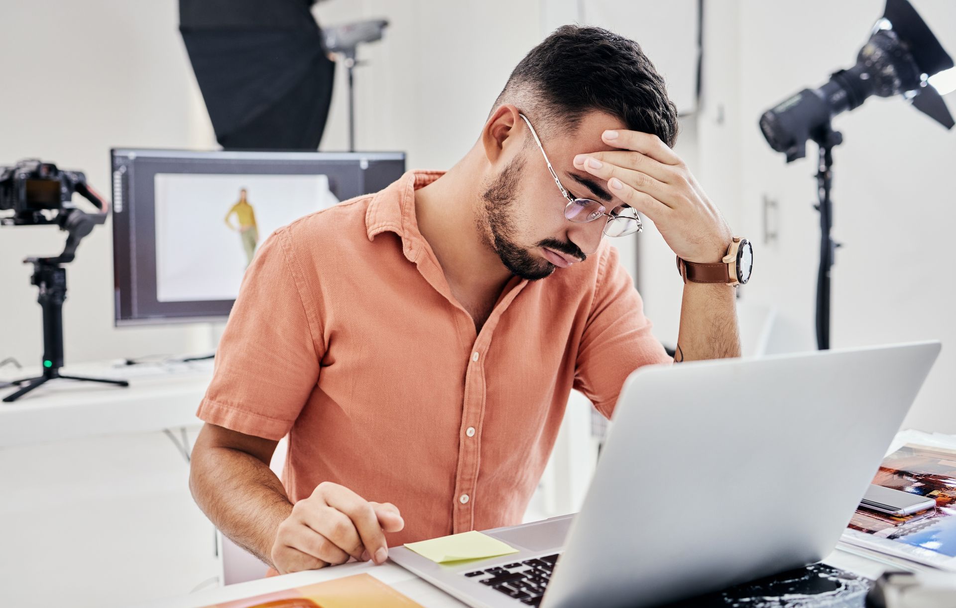 Stressed man in an orange shirt leaning over a laptop in a bright office, holding his forehead.