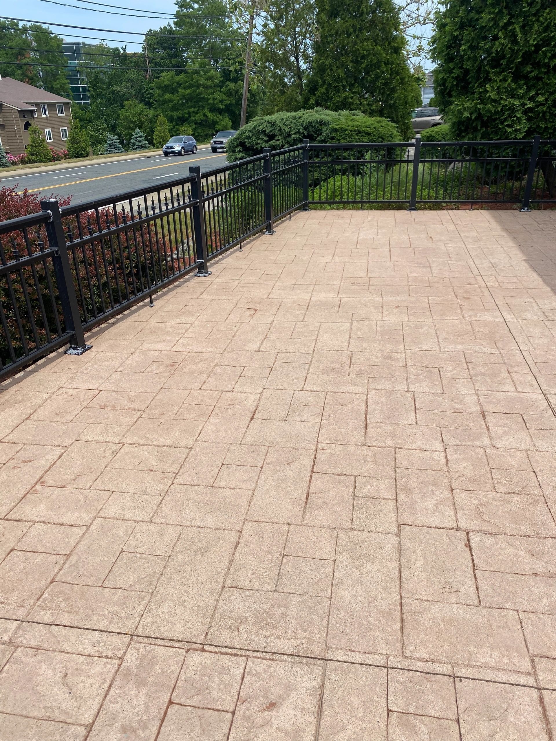 Empty brick patio with black railing beside a road and greenery under a sunny sky