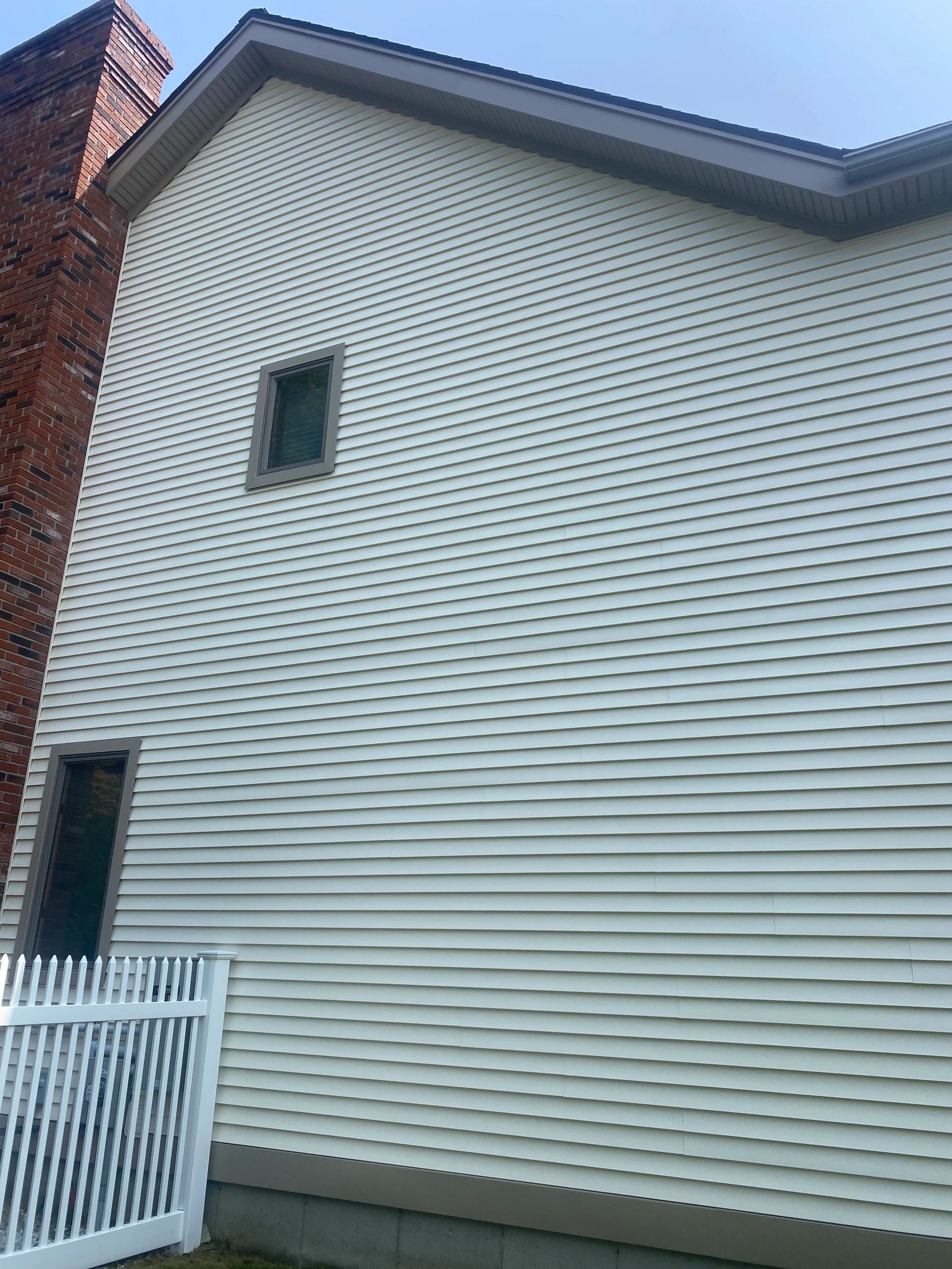 White vinyl-sided house exterior with a small window, red brick chimney, and white fence.