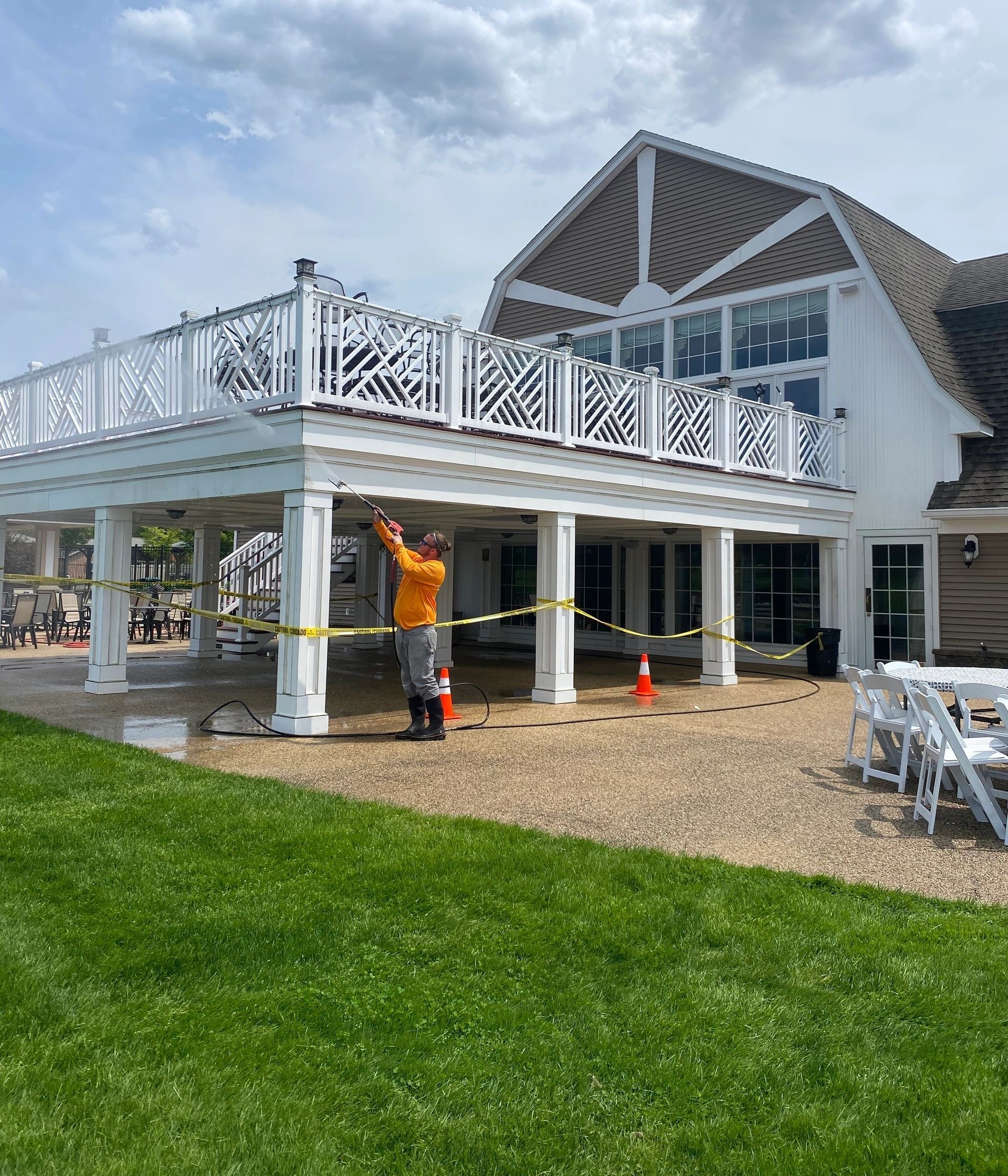 House with white deck under construction, worker in orange vest, caution tape, and green lawn under cloudy sky
