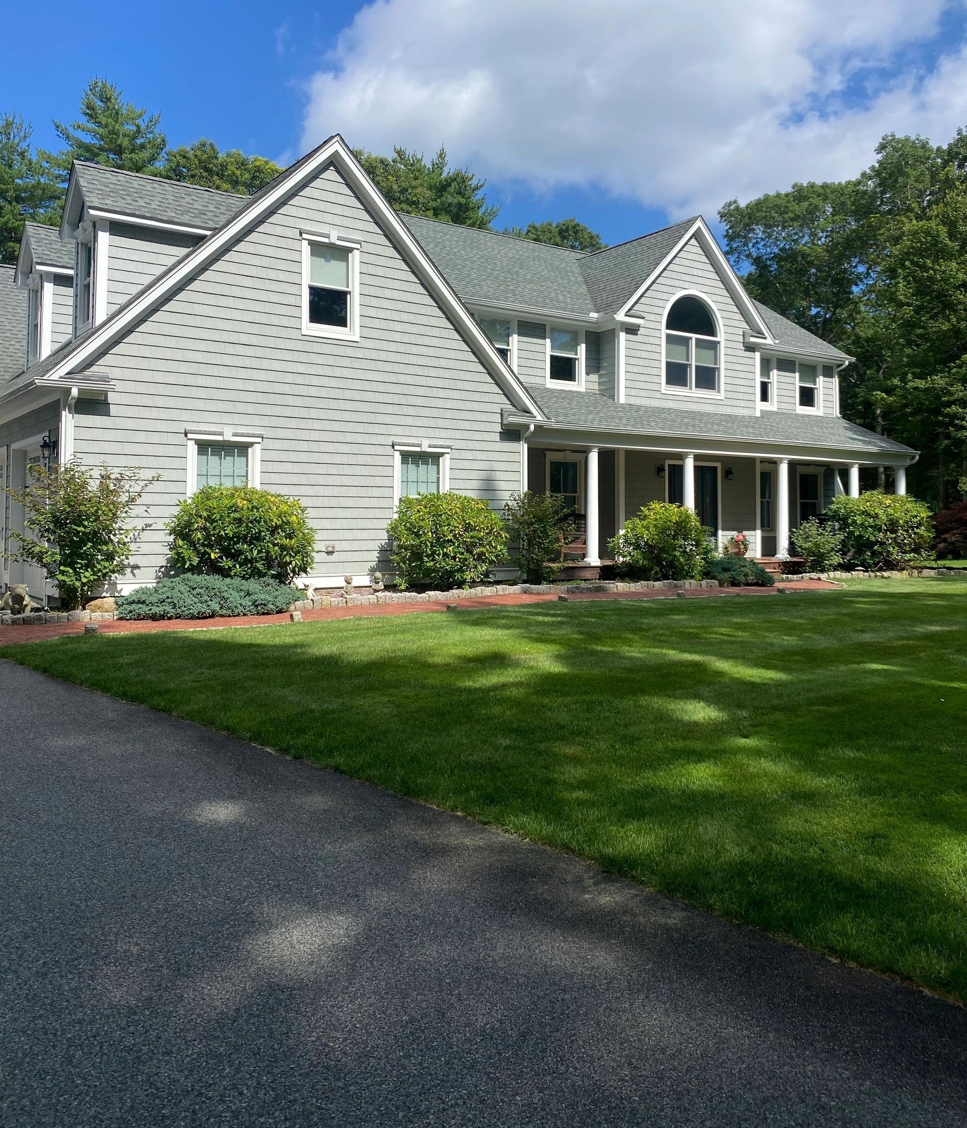 White shingled house with front porch, dormers, and green lawn beside a gravel driveway under a blue sky