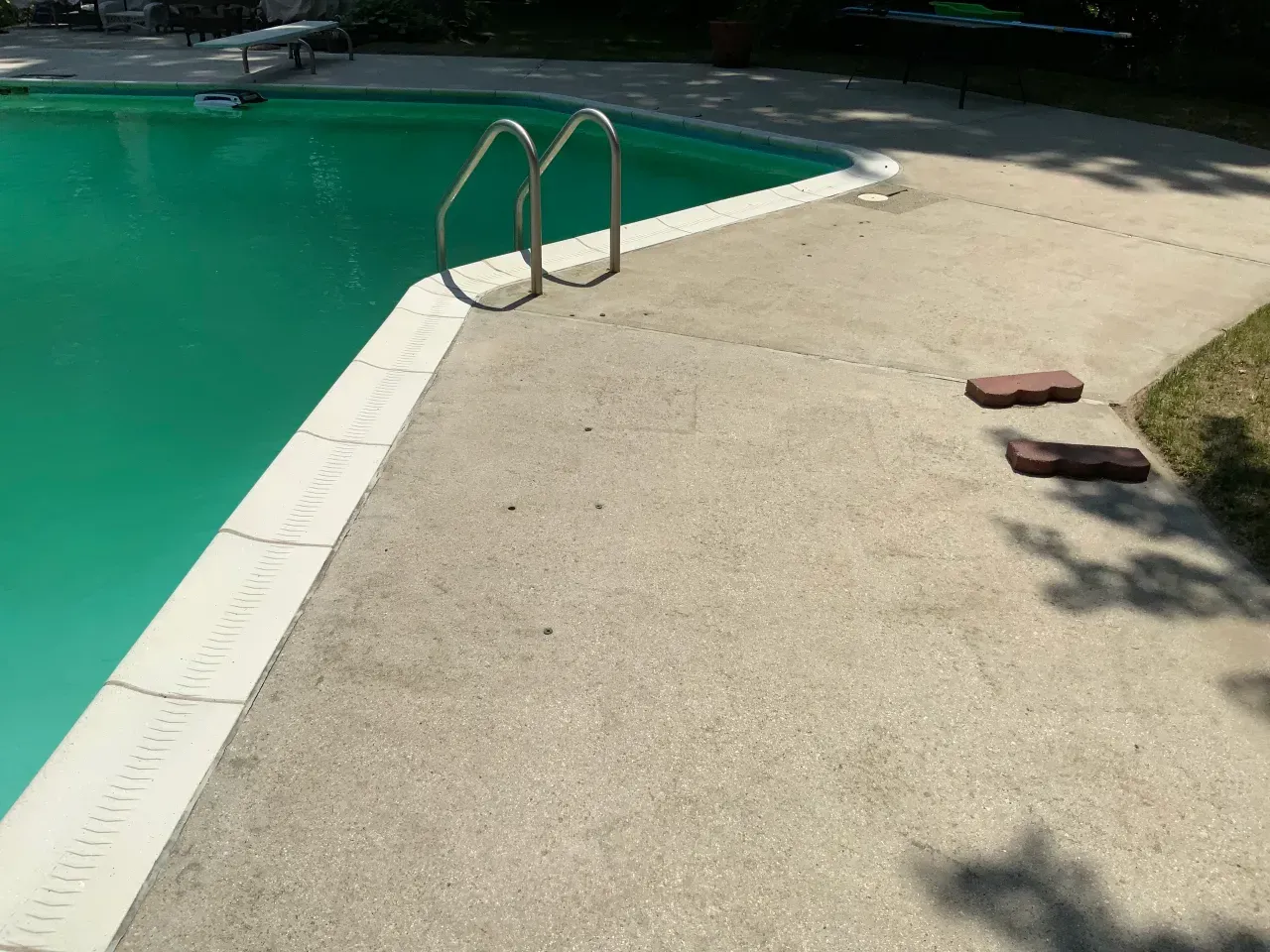 Poolside concrete deck beside green swimming pool with a metal ladder and two bricks on the ground