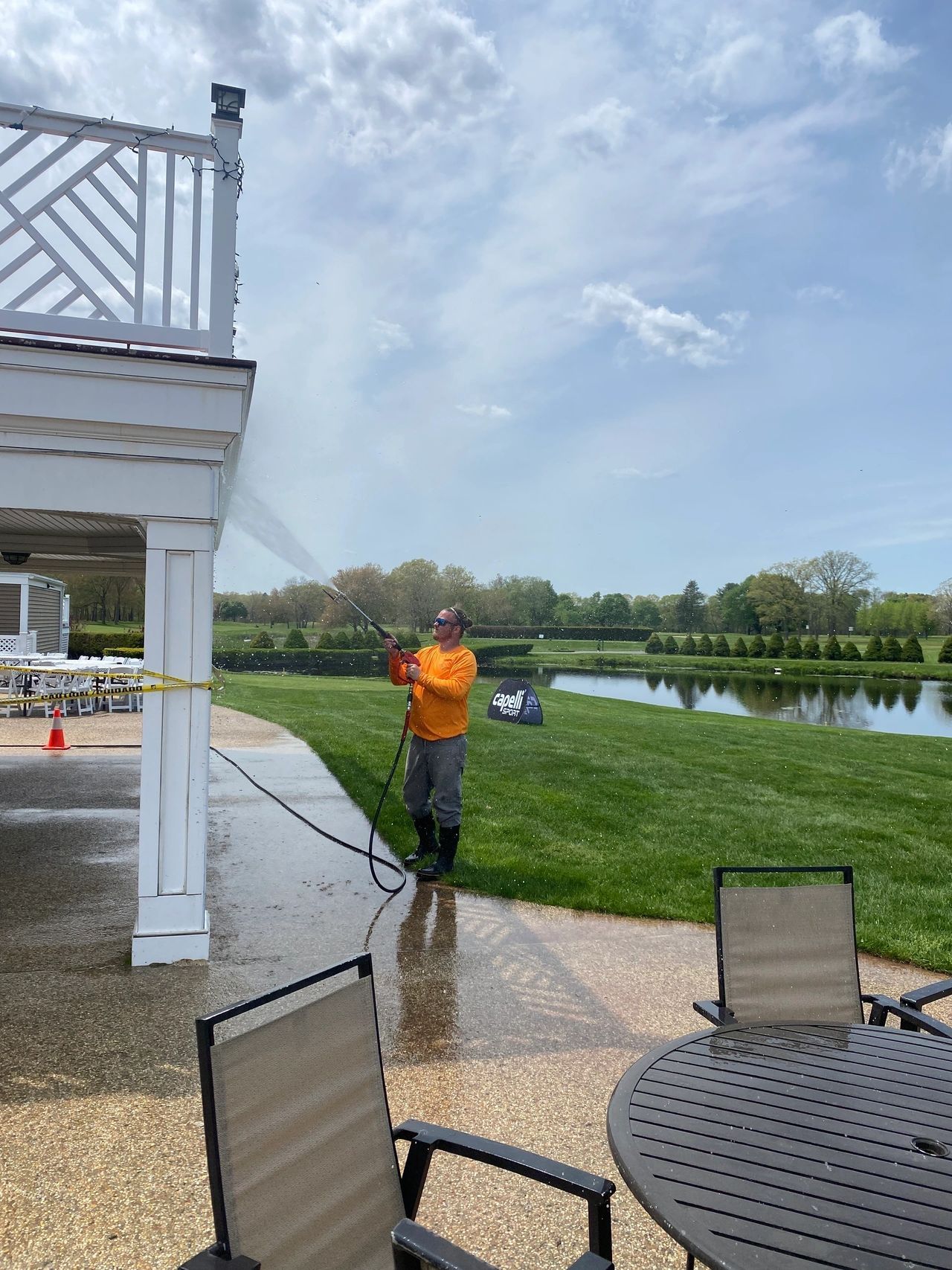 Person spraying water on a patio beside a white house, overlooking a lake and grassy yard.