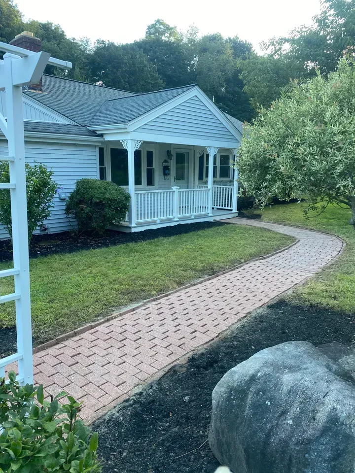White house with porch beside a brick path curving through a green yard, with trees and rocks nearby.