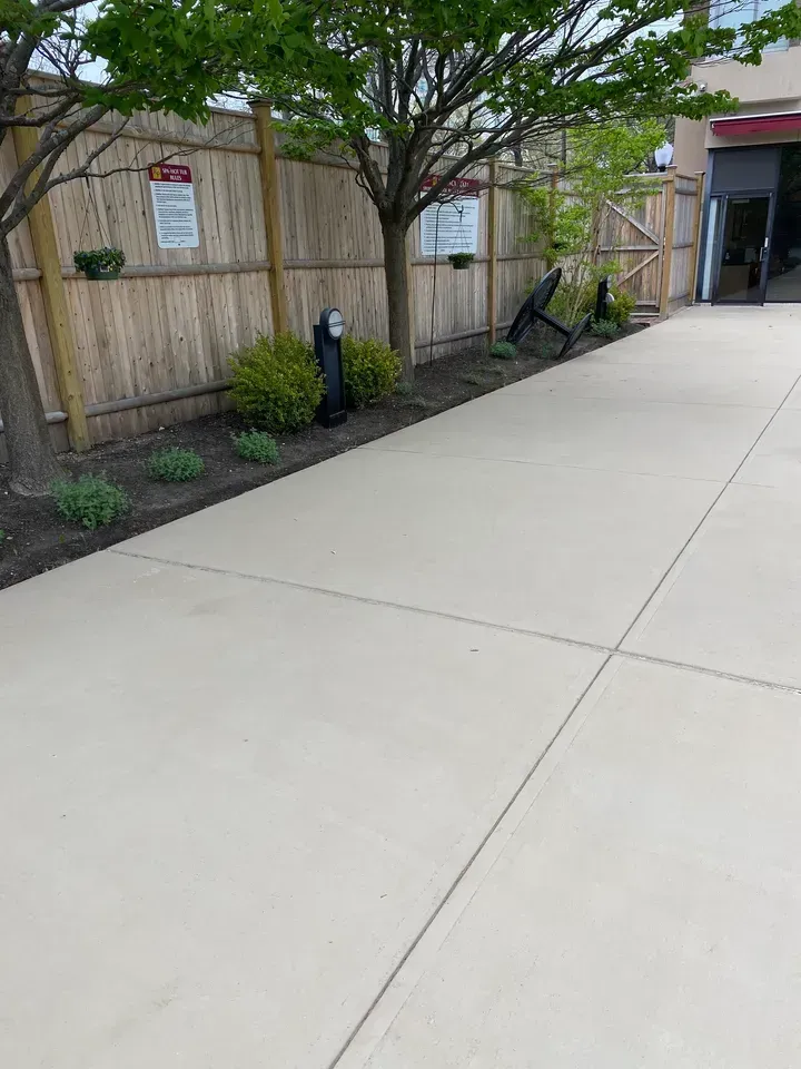 Concrete walkway beside a wooden fence and trees, with a building entrance in the background.