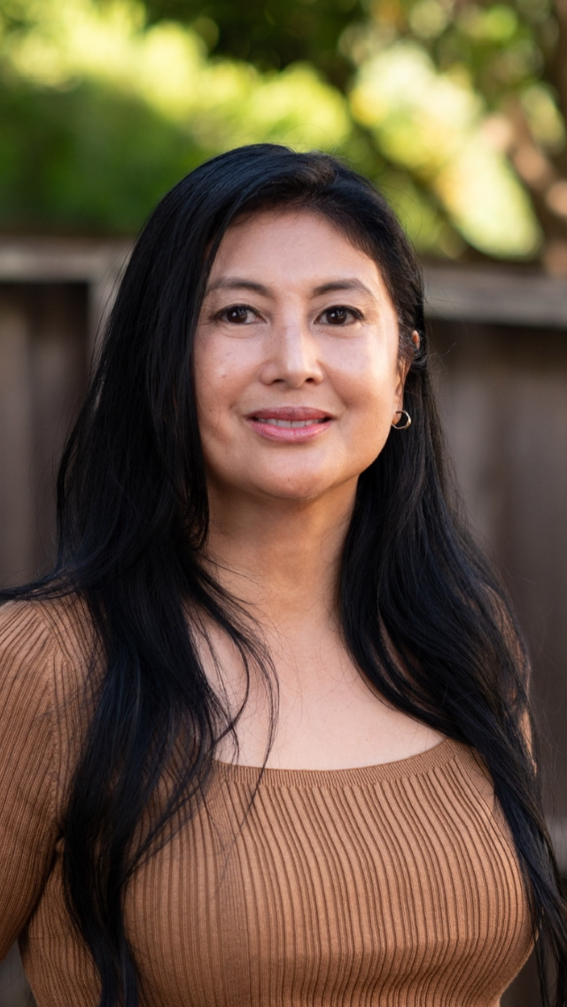 A woman with long black hair is standing in front of a wooden fence.