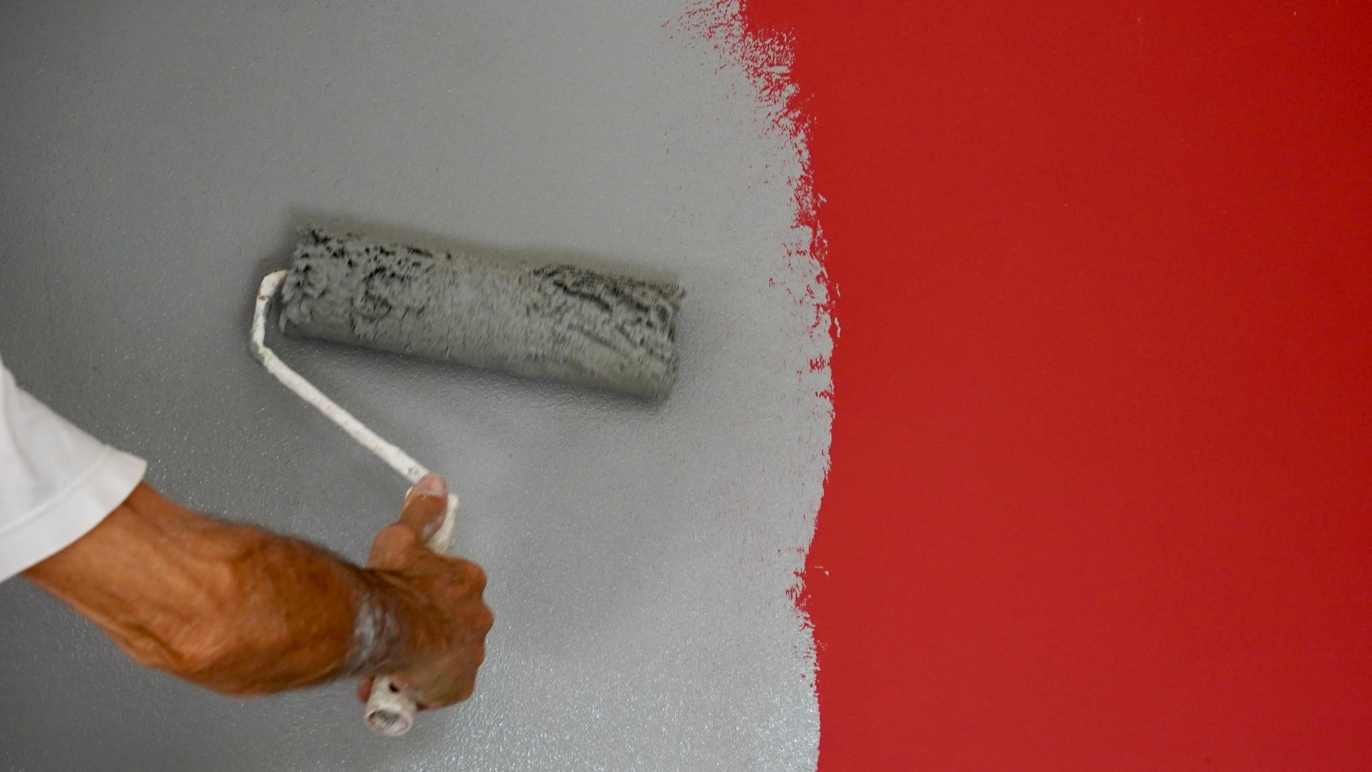 A hand paints a wall gray beside a bright red section with a paint roller.