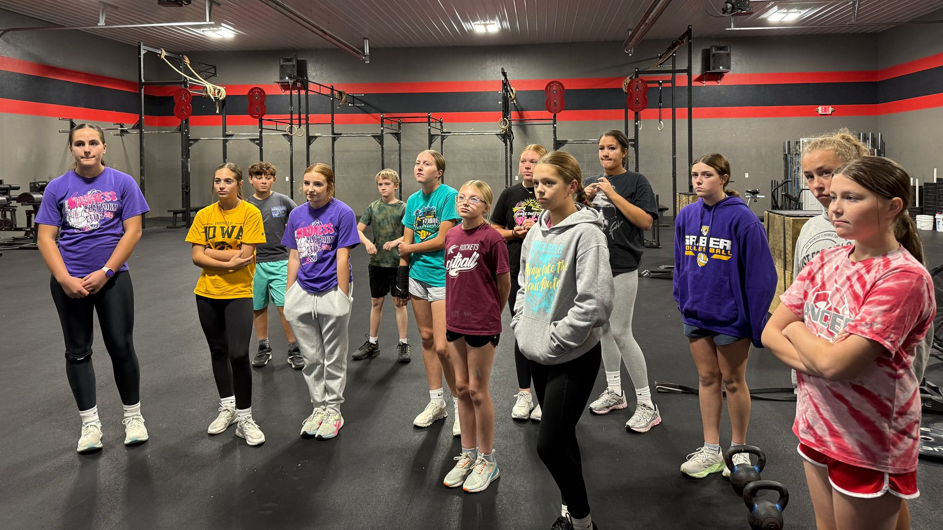 A coach instructs a group of people in a gym as they hold wooden dowels and practice squat form.