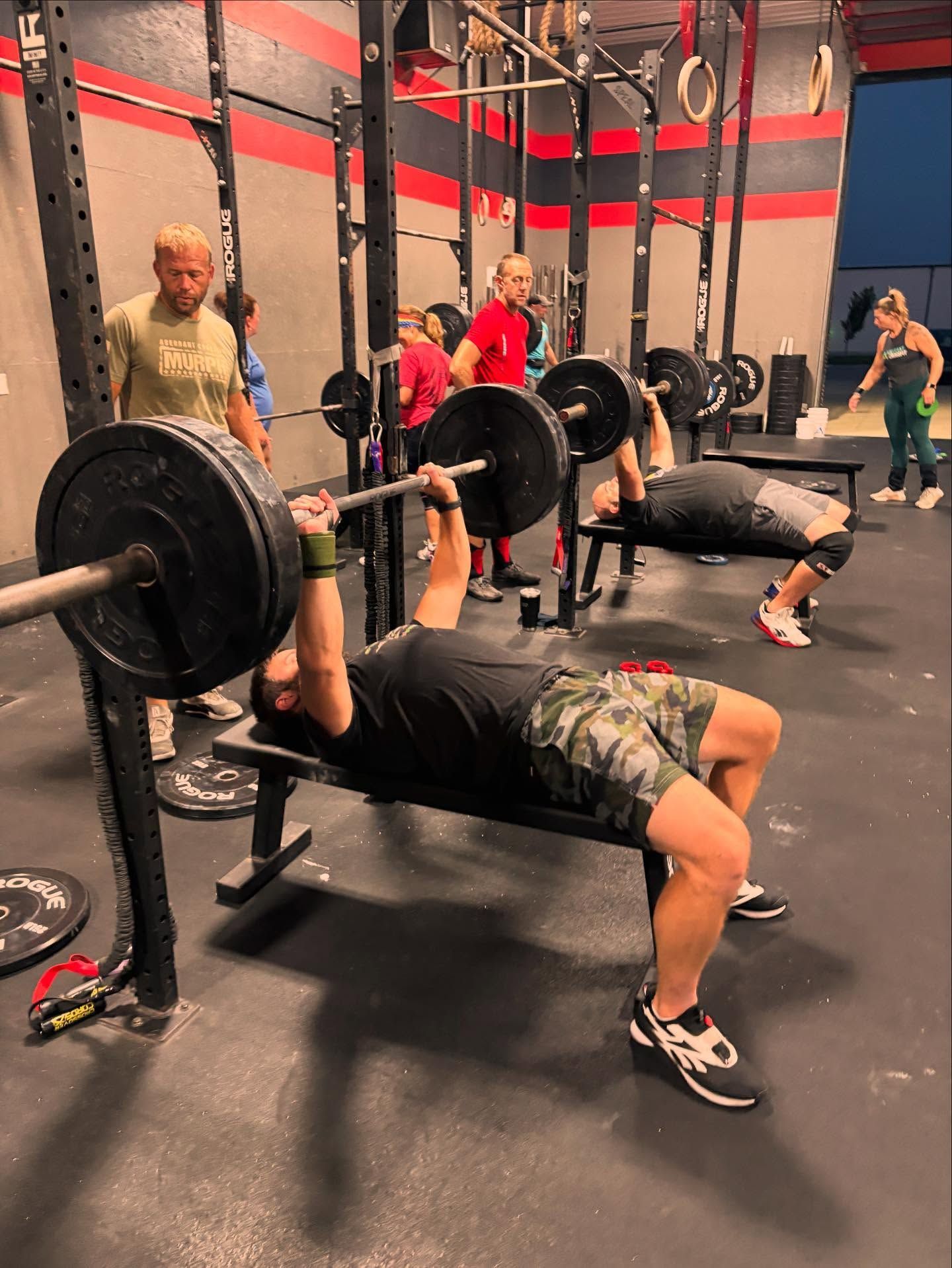 People exercise in a gym with weight racks; two people perform barbell bench presses while others stand nearby.