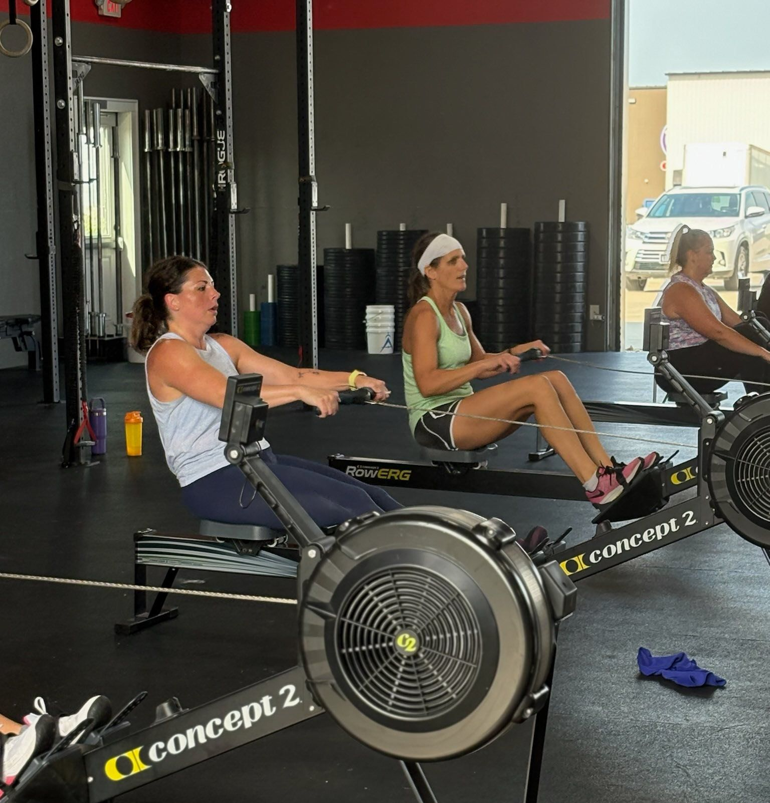 Three people exercise on indoor rowing machines in a gym with weights in the background.