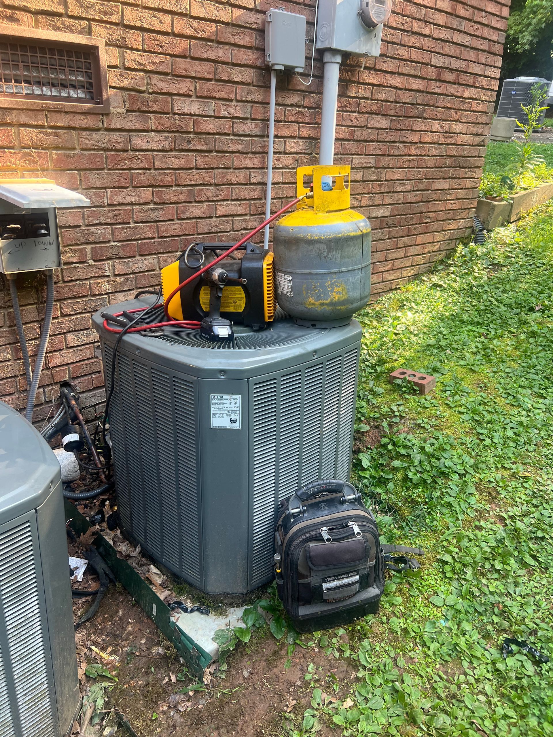 HVAC unit with tools, near a brick building. A yellow tank and black pump are on top. Black bag on the ground.