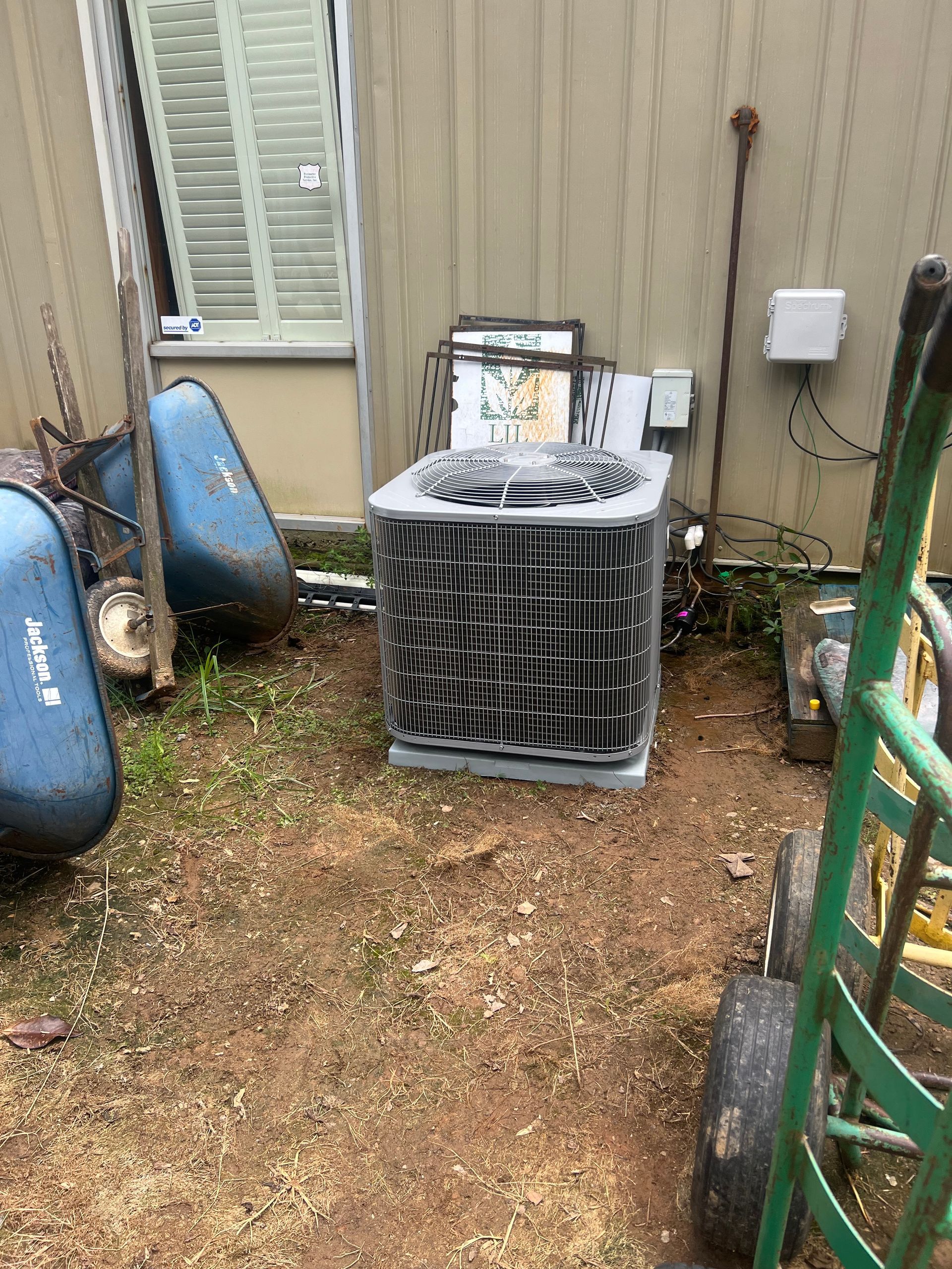 Air conditioner unit outside near a building. Surrounded by yard tools and brown dirt.