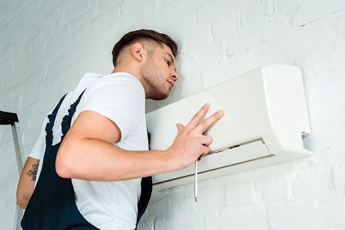 Man in overalls examines a white wall-mounted air conditioner unit. White brick wall background.