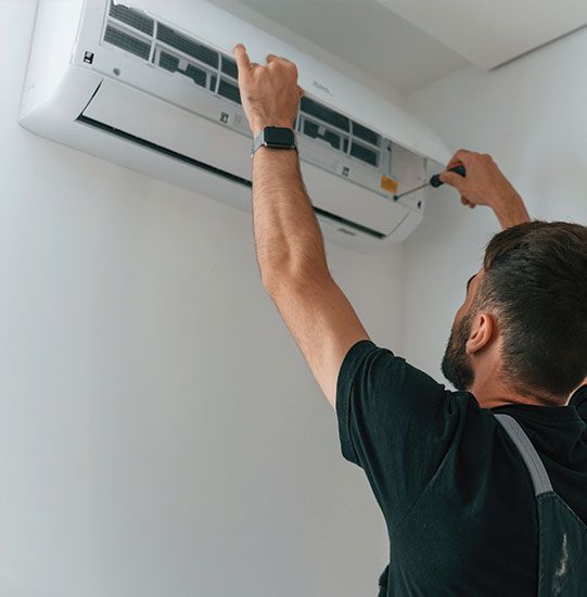 A man in a black shirt repairs a wall-mounted air conditioner with a screwdriver. White room.