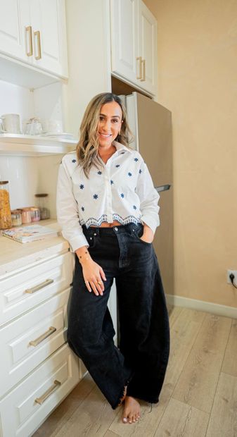 Woman in white blouse and dark jeans leaning in a bright kitchen beside white cabinets and a refrigerator