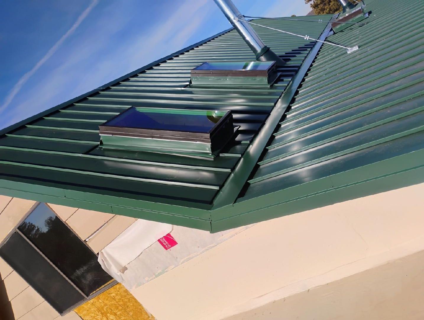 Green metal roof with skylights and chimney against a blue sky.
