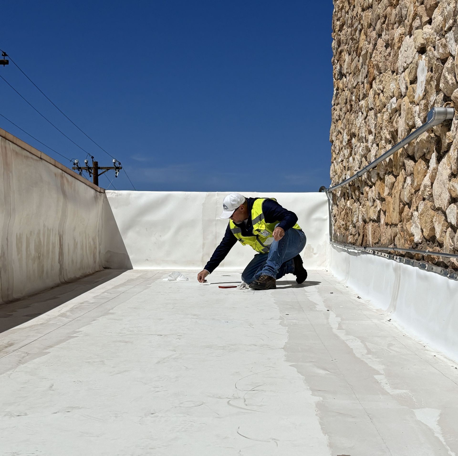 A worker kneels on a flat roof, inspecting it. White roof, stone wall, bright blue sky.