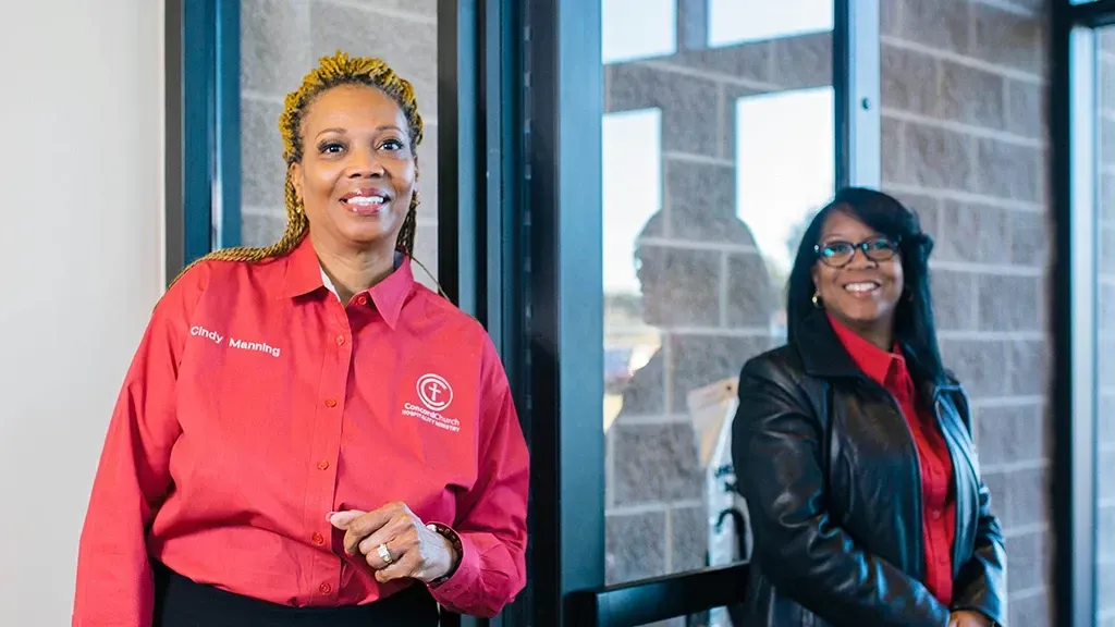 Two women smiling, one in red shirt, the other in black jacket, in an office setting.