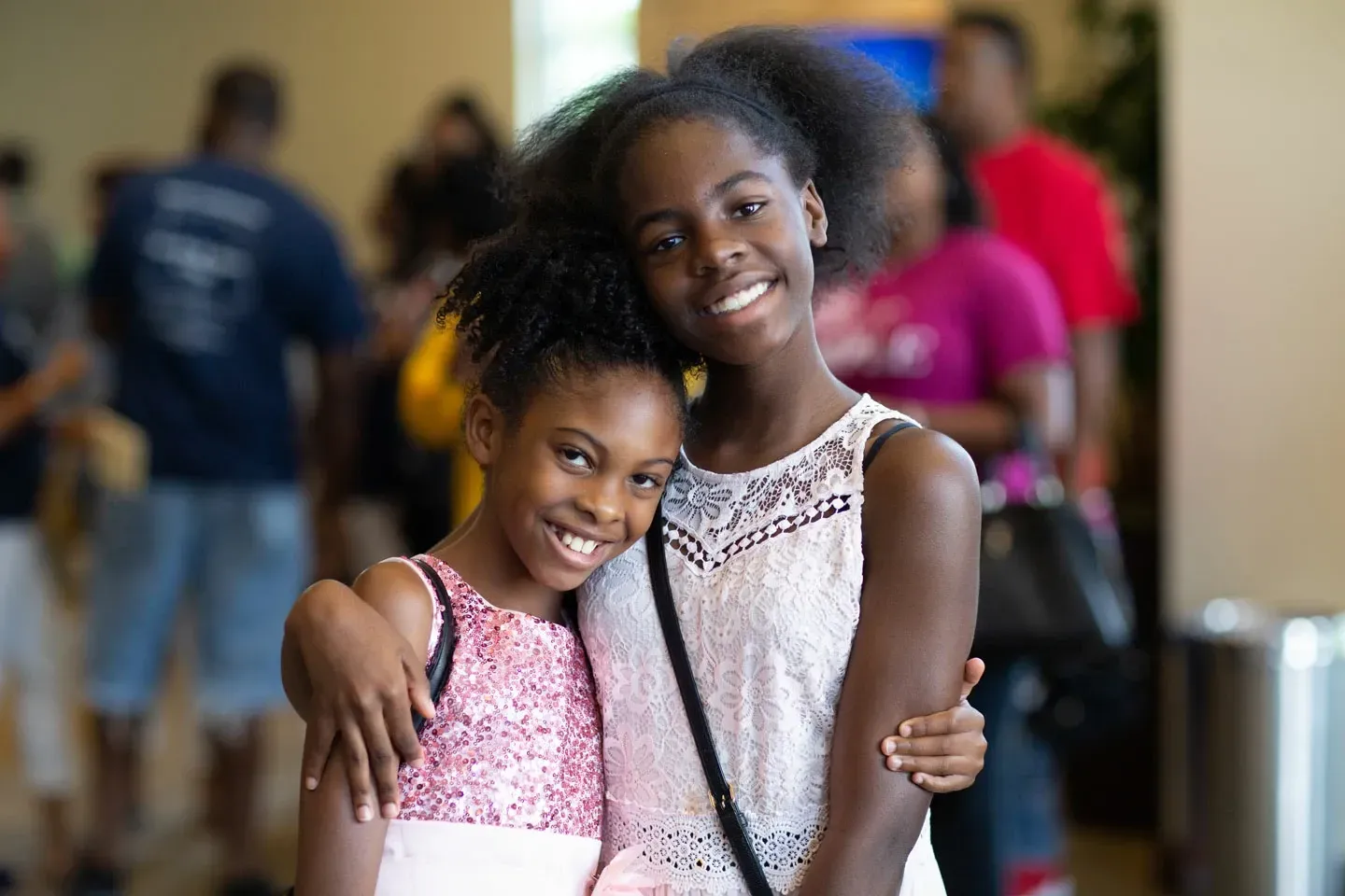 Two smiling Black girls embrace indoors, one in a white top, the other in pink, with blurred people in the background.