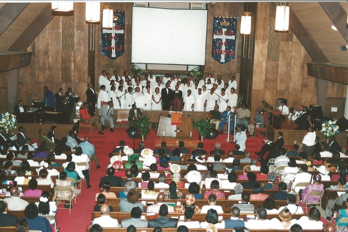 Church service: Choir in white robes stands before congregation. Red carpet, wooden paneling, banners.
