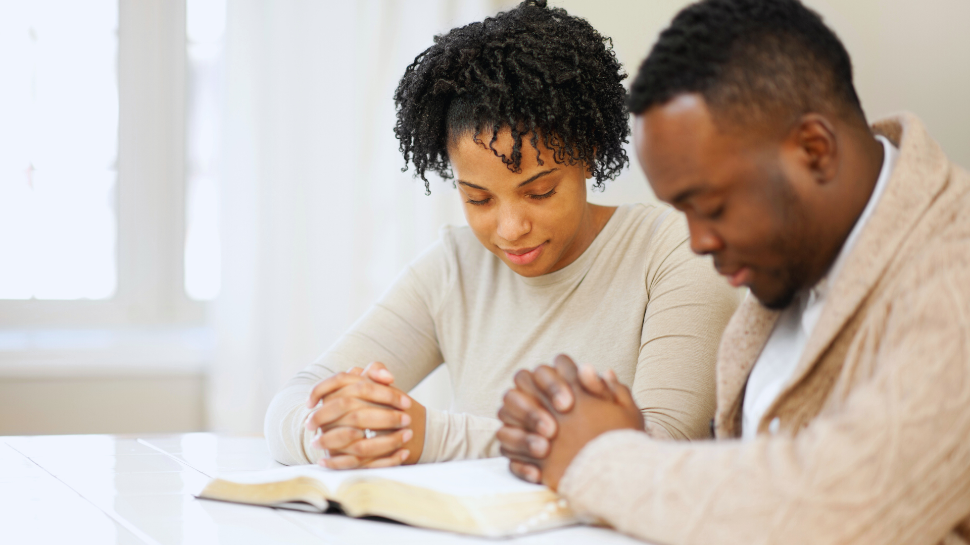 A Black couple with hands clasped, heads bowed over an open book, praying together at a table.