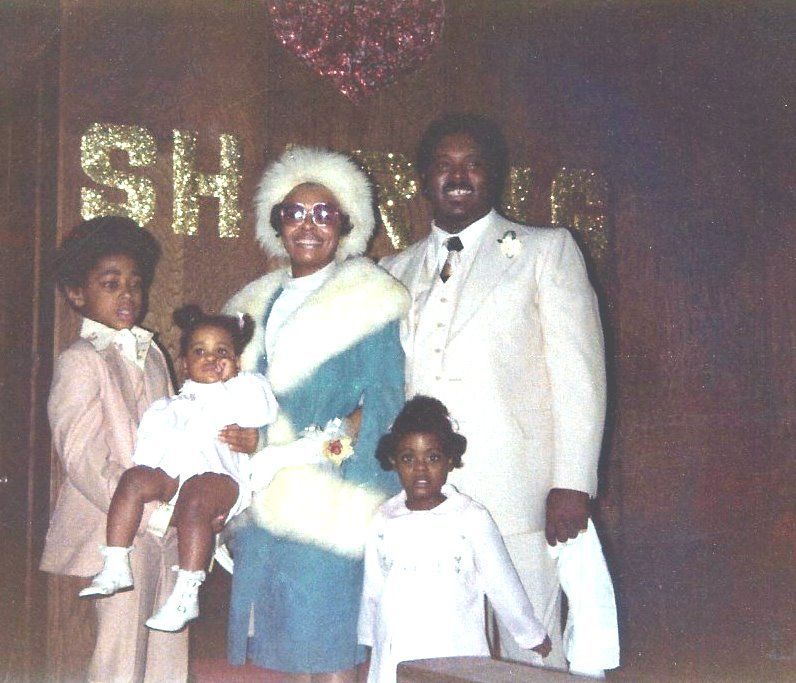 Family posing for photo at a wedding shower. Woman in fur coat, man in white suit, children in formal attire.