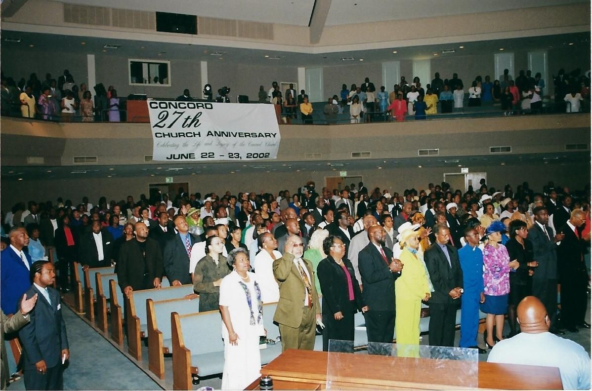 Large gathering in a church for the 27th Anniversary. People stand, mostly African American.