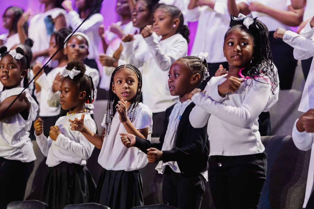 Children's choir performing. They wear white tops, some with black skirts/pants, waving hands. Pink and purple backdrop.