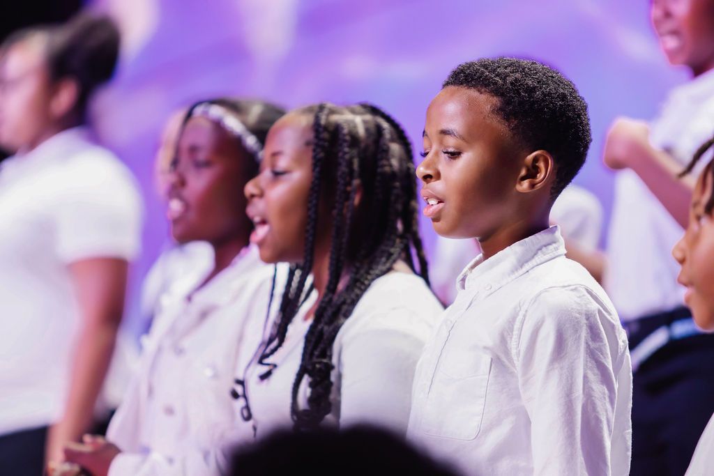 Children's choir performing on stage, dressed in white shirts, singing with focused expressions. Purple background.