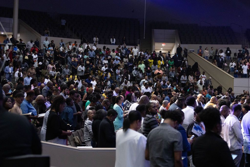 Large gathering in an auditorium; many people stand and sit, looking forward, in various clothing colors.