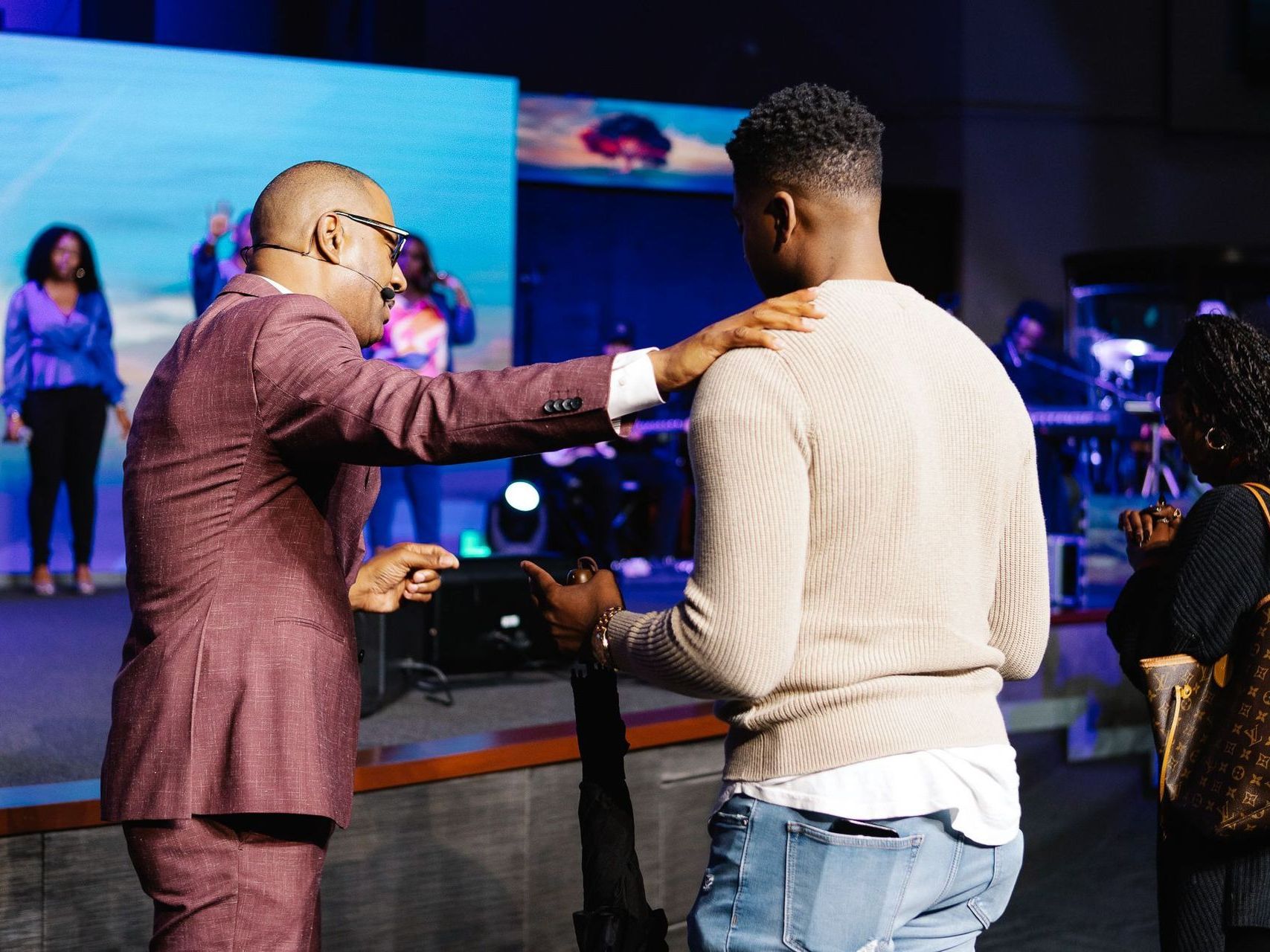 A man in a suit blesses another man on stage. A band plays behind them.