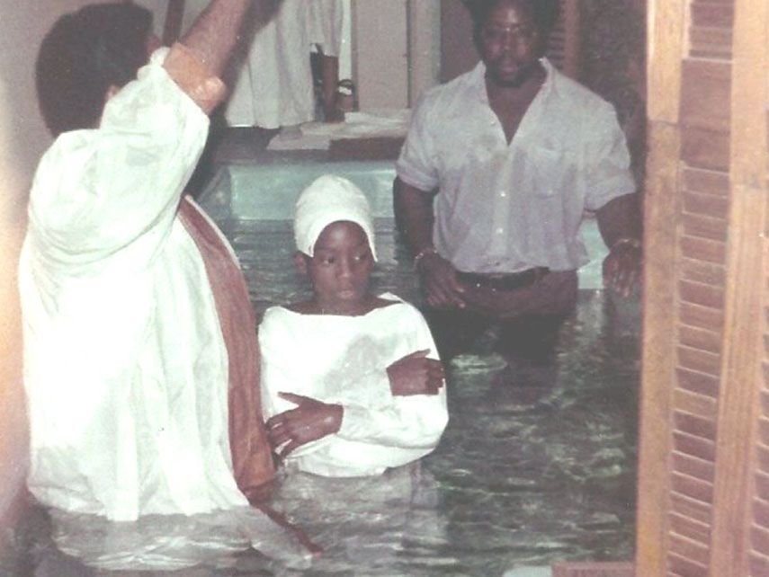 People in waist-deep water: one woman in a headscarf and two men, one raising a hand. Likely a baptism.