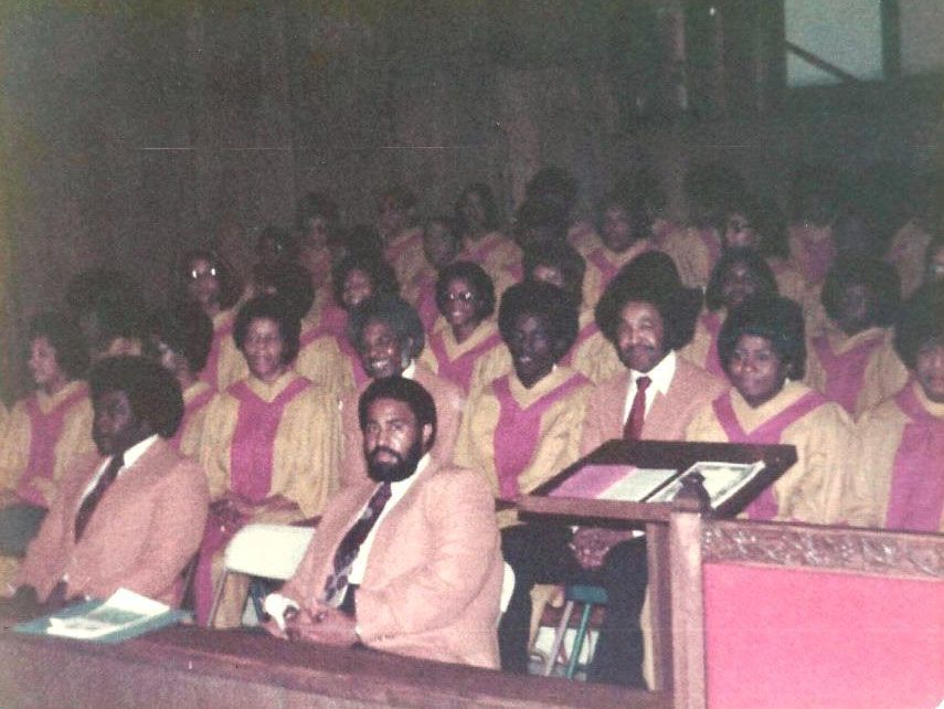Choir in gold robes with pink trim, singing in a church. A man in a suit sits in front.
