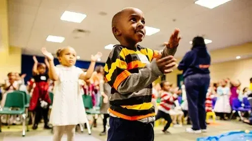 Children clapping in a brightly lit room; a boy in a striped shirt and a girl in a white dress are in focus.