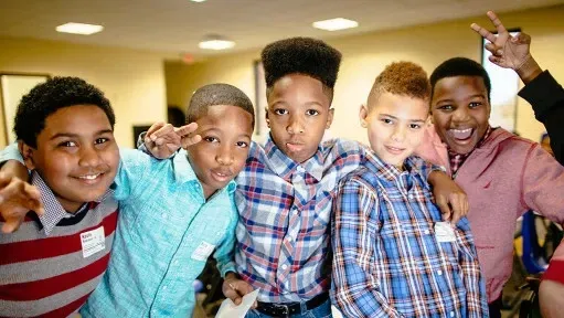 Group of smiling boys, arms around each other, posing for a photo. Bright, colorful shirts and diverse hairstyles.