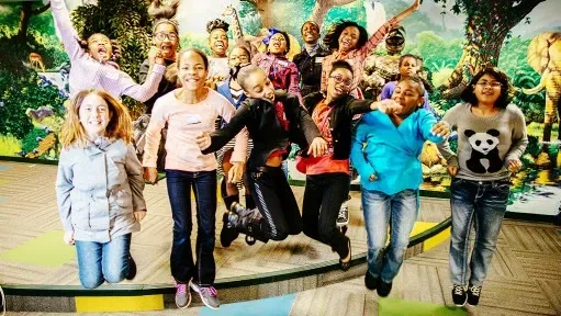 Group of diverse kids jumping and smiling indoors near a colorful jungle-themed backdrop.