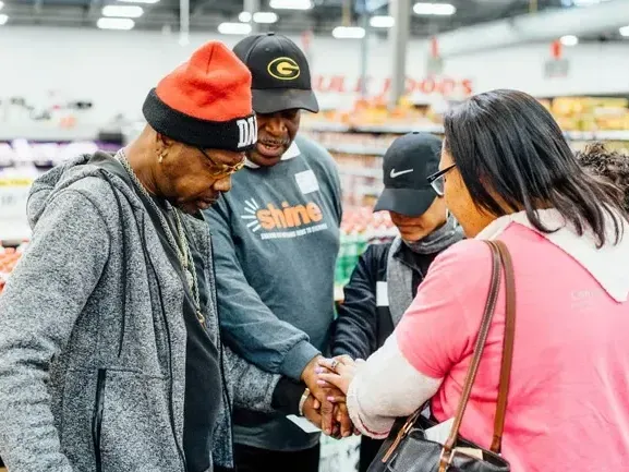 Four people with hands together in a grocery store; praying.