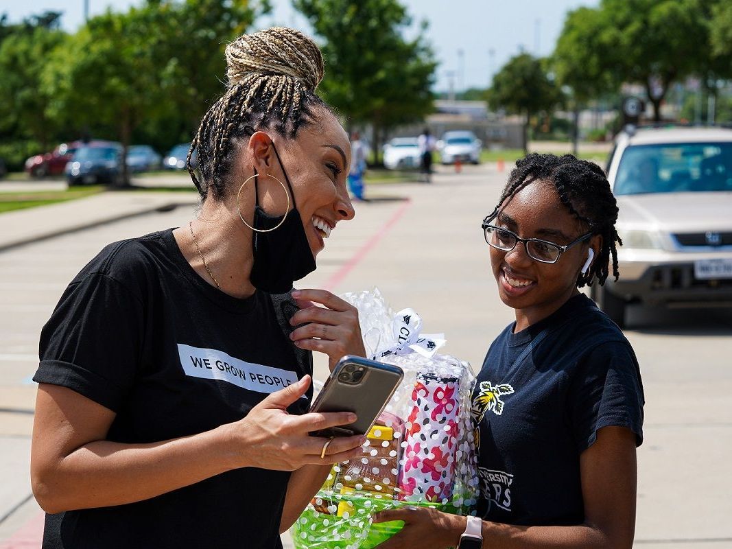 Woman and girl laugh, holding a phone and gifts outdoors. Both wear masks and dark shirts.