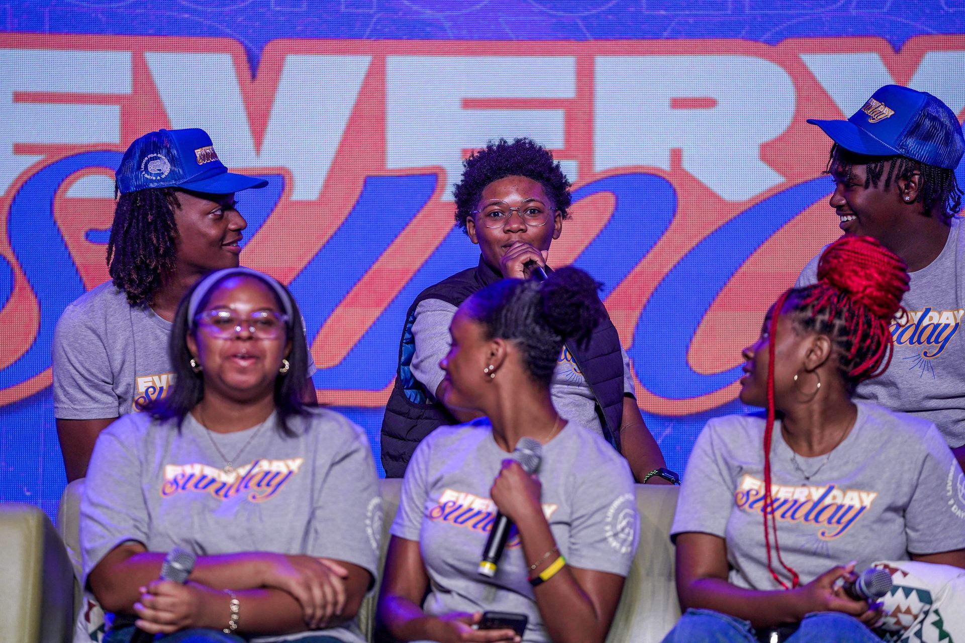Group of people on stage wearing matching shirts and hats with a logo. They are holding microphones and looking towards the center.