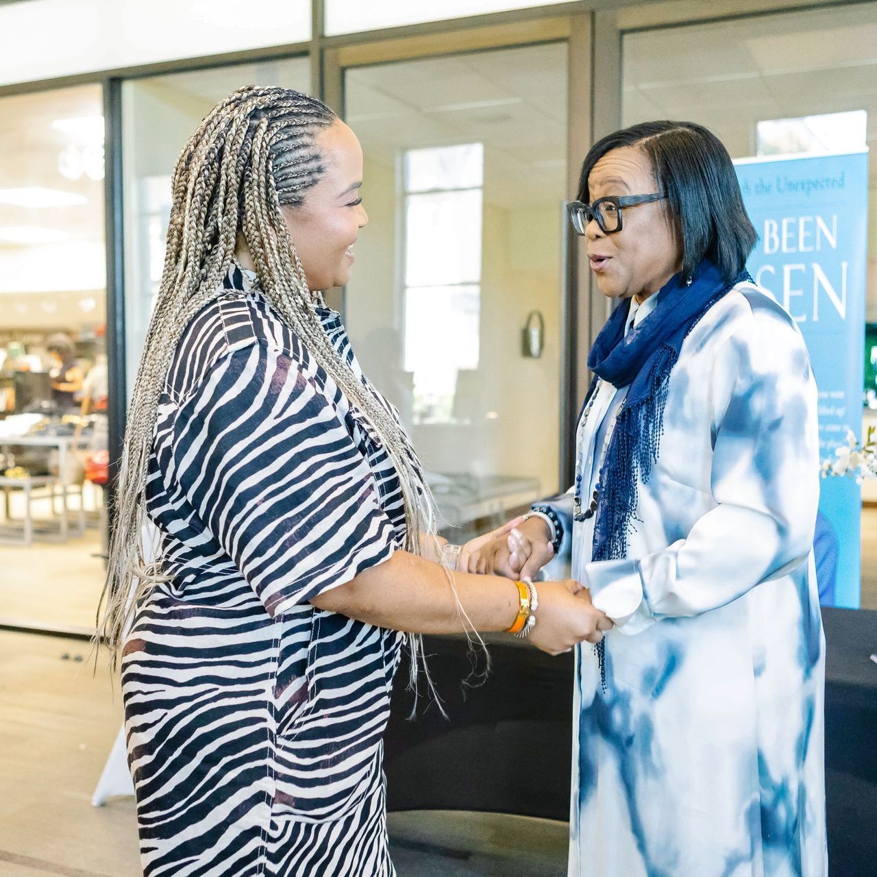 Two women smiling and shaking hands in a well-lit room. One in zebra-print, the other in a blue tie-dye dress.